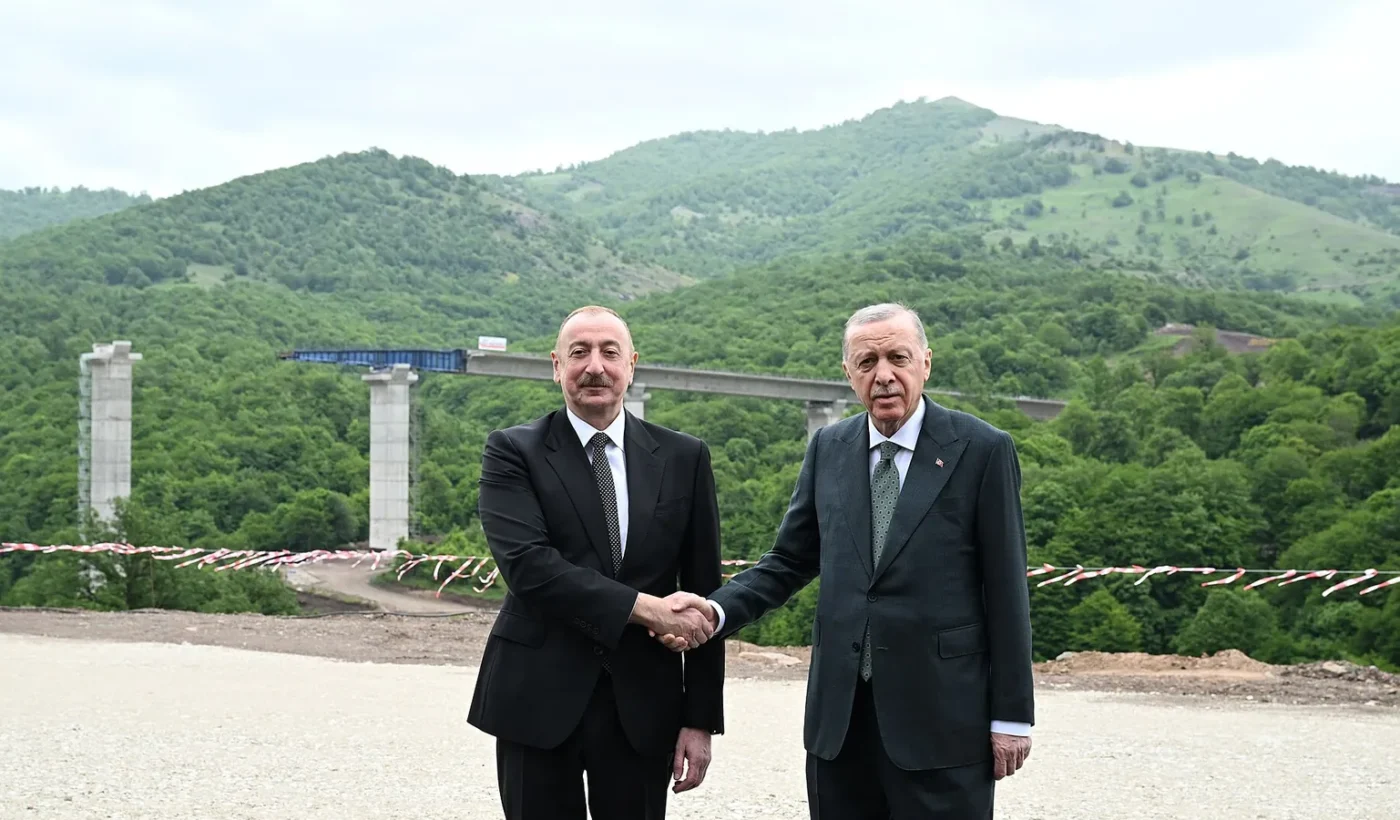 Ilham Aliyev and Recep Tayyip Erdogan shaking hands in front of a highway bridge in a mountainous area