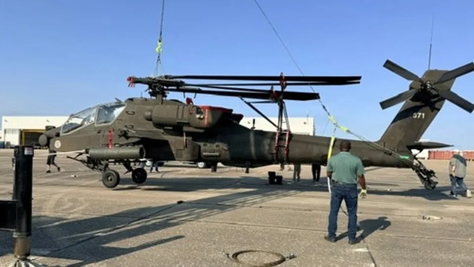 An AH-64E Apache helicopter with folded main rotor blades hangs from a crane on an airfield during lifting tests, with ground crew standing nearby