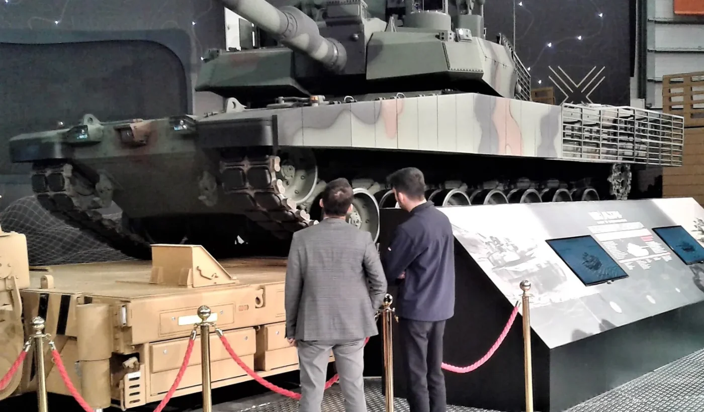 A camouflaged Turkish Altay main battle tank at an exhibition with two visitors in front of it