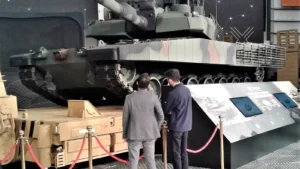 A camouflaged Turkish Altay main battle tank at an exhibition with two visitors in front of it
