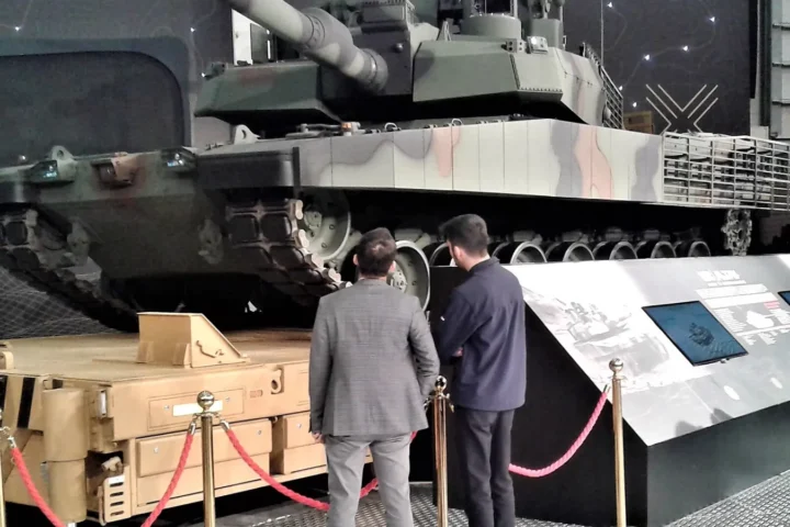A camouflaged Turkish Altay main battle tank at an exhibition with two visitors in front of it