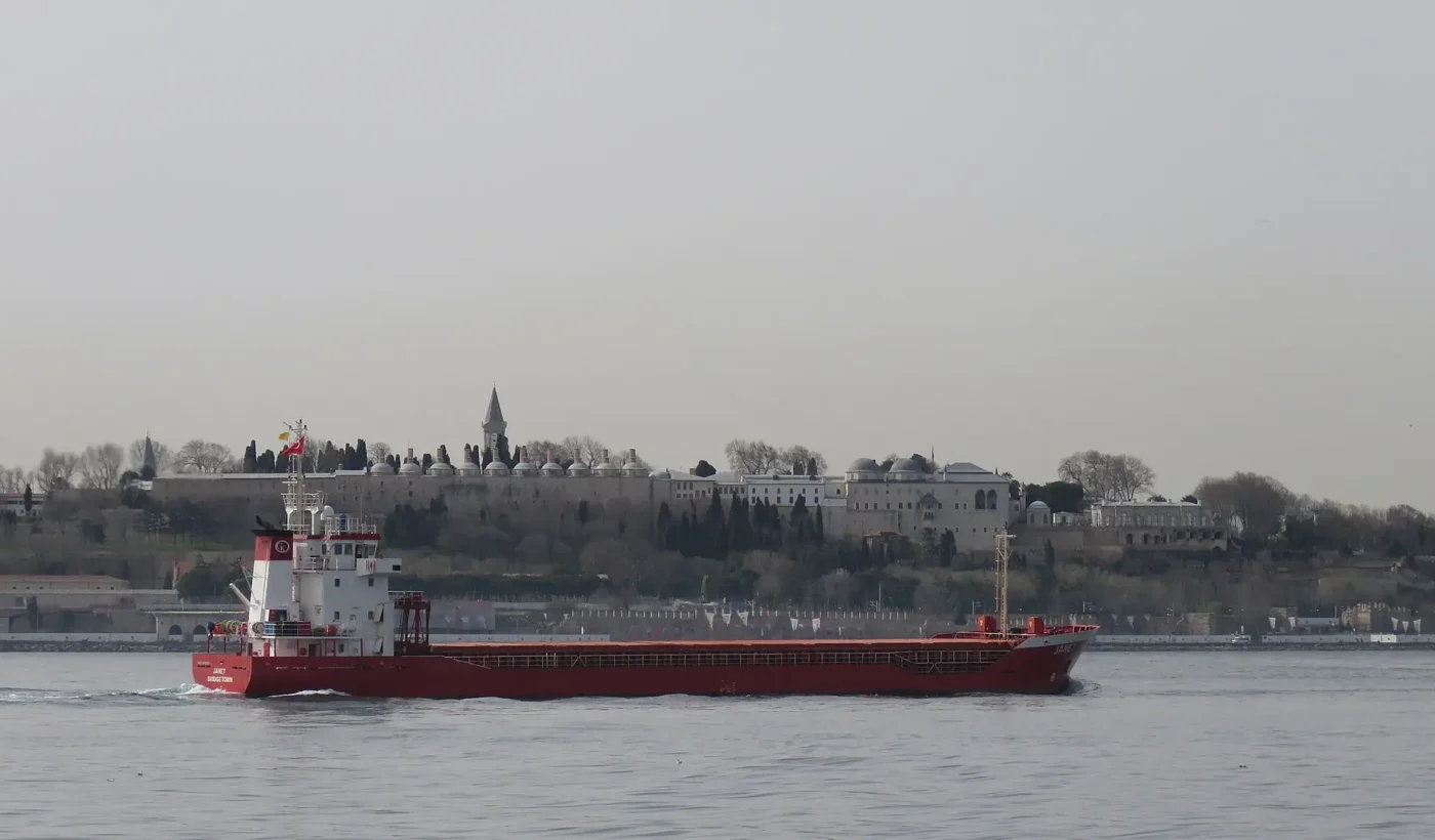 A red-hulled general cargo ship sails along the Bosphorus with the buildings of Topkapı Palace on the shore behind it