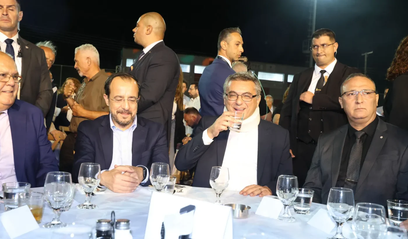 Two men in suits seated at a table among other guests at an outdoor festival in a village