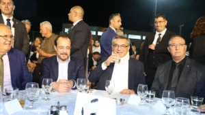 Two men in suits seated at a table among other guests at an outdoor festival in a village