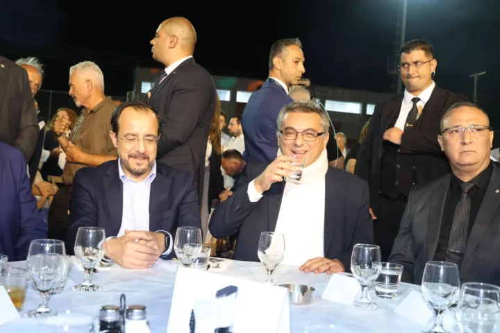 Two men in suits seated at a table among other guests at an outdoor festival in a village