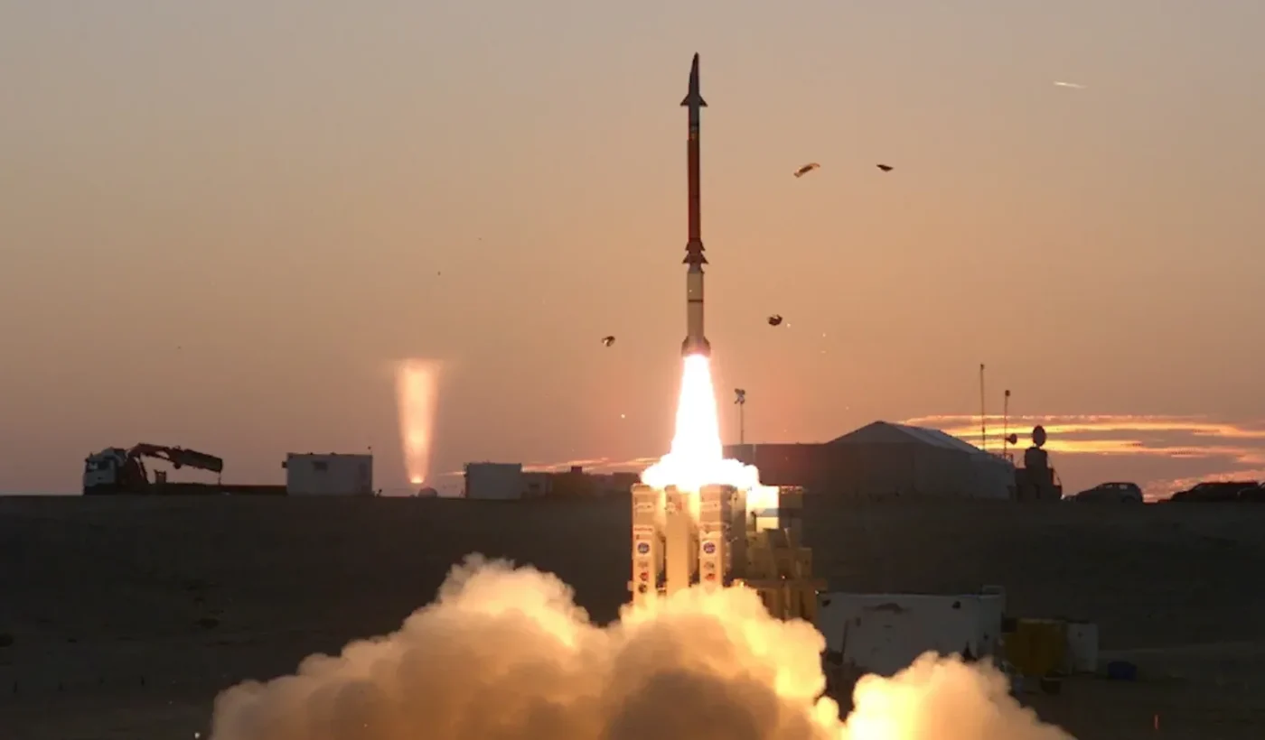 A David’s Sling Stunner interceptor rising from its launcher in a plume of fire and smoke at sunset in the Israeli desert