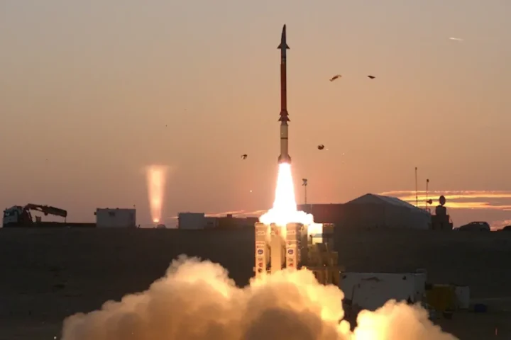 A David’s Sling Stunner interceptor rising from its launcher in a plume of fire and smoke at sunset in the Israeli desert