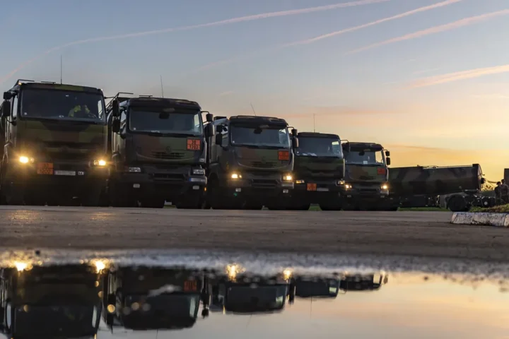 Row of French military fuel tanker trucks with headlights on at dusk, reflected in a puddle