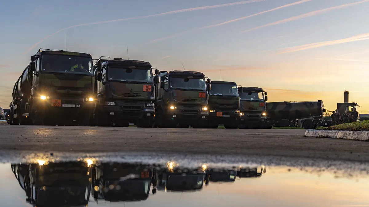 Row of French military fuel tanker trucks with headlights on at dusk, reflected in a puddle