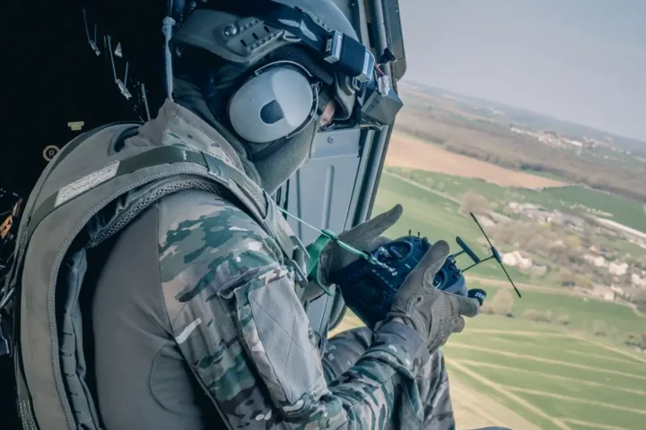 A French soldier in a helicopter doorway holding a drone controller with a headset-mounted video receiver, over farmland