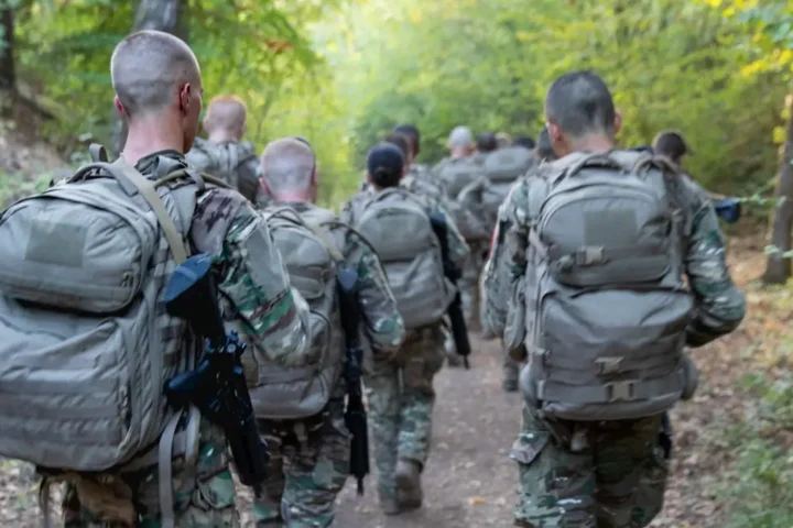 Young French national service recruits in camouflage with backpacks marching in a column along a forest path