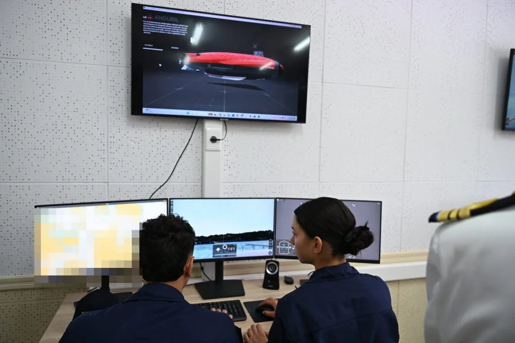 Two trainees in front of three simulator screens, with a television above showing a red underwater vehicle
