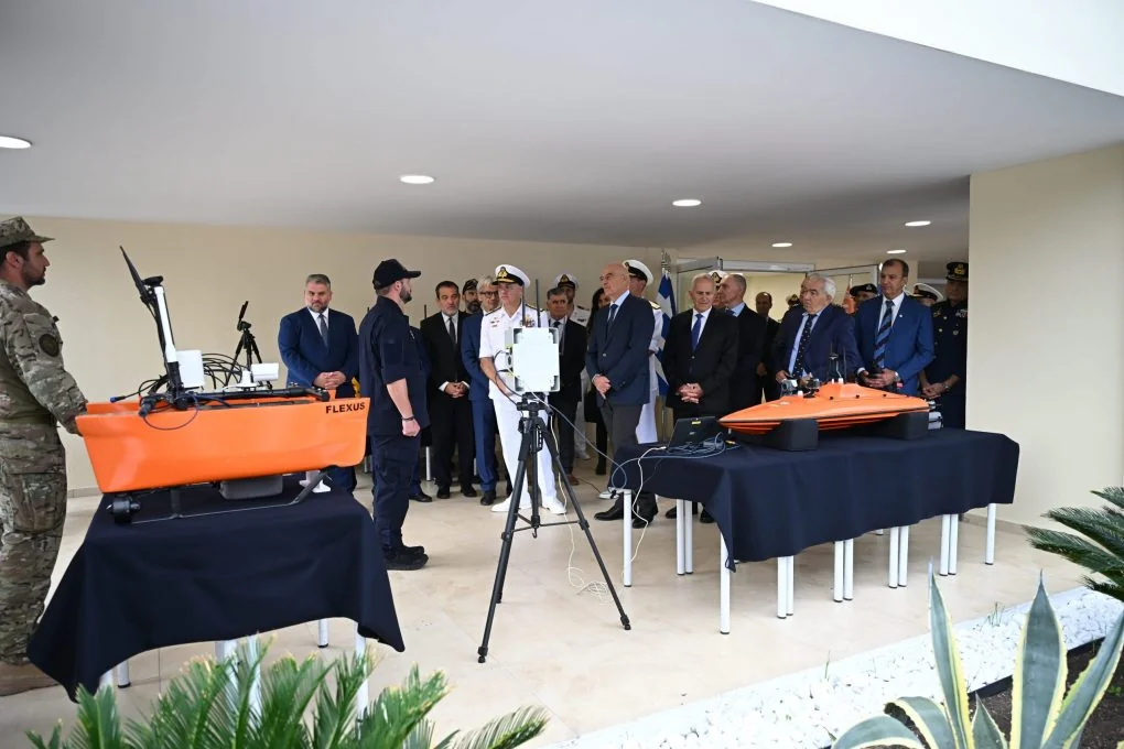 Nikos Dendias and officers watching a presentation of small orange unmanned vessels on tables