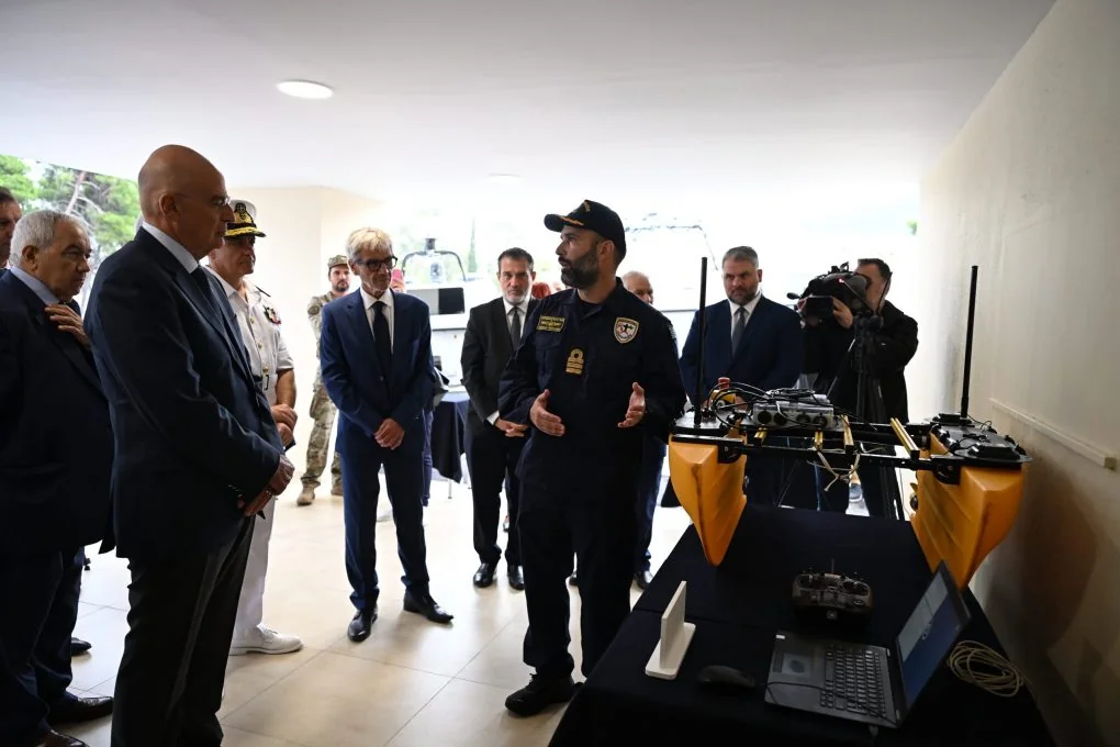 An officer explaining to Nikos Dendias the operation of a small yellow twin-hull vessel with antennas, next to a laptop and a controller