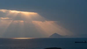 A merchant ship on the horizon of the Strait of Hormuz at dusk, with sunbeams breaking through clouds over a headland