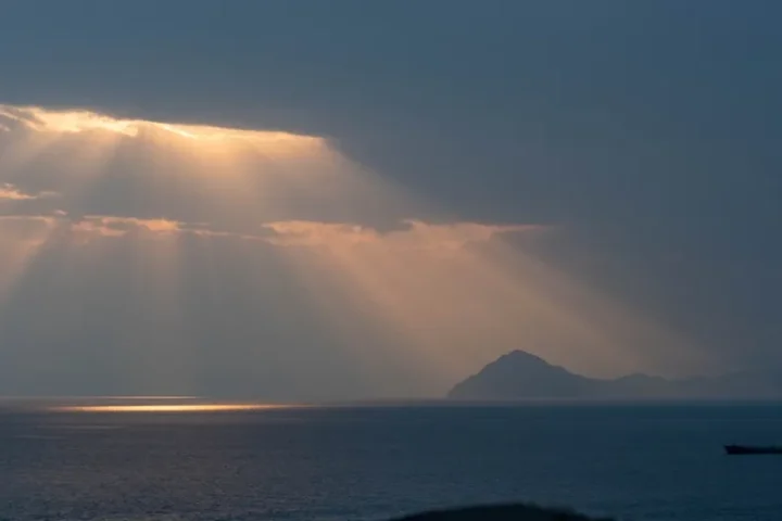 A merchant ship on the horizon of the Strait of Hormuz at dusk, with sunbeams breaking through clouds over a headland