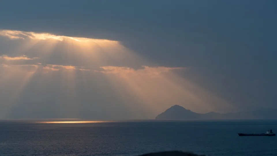 A merchant ship on the horizon of the Strait of Hormuz at dusk, with sunbeams breaking through clouds over a headland
