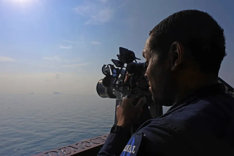 A U.S. Navy sailor uses a sextant on the bridge wing of a destroyer during a Strait of Hormuz transit, with low islands on the hazy horizon