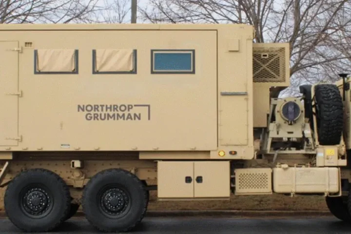 Tan shelter van with the Northrop Grumman logo mounted on a military truck, shown with its sides closed