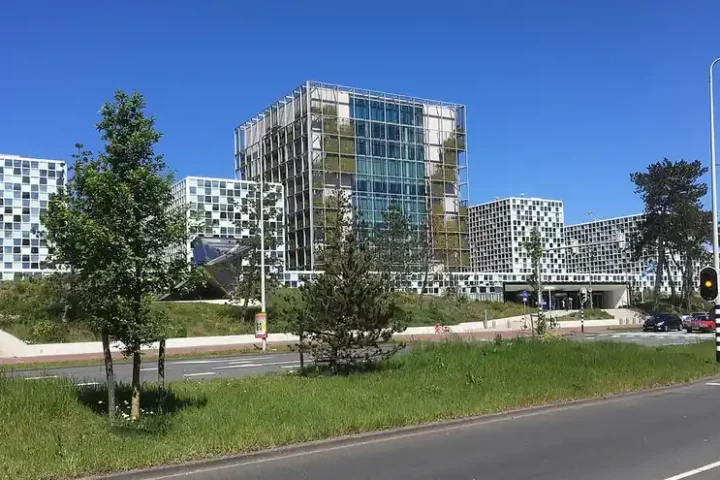 The glass and green-facade main tower of the International Criminal Court building in The Hague, with white and blue office blocks beside it
