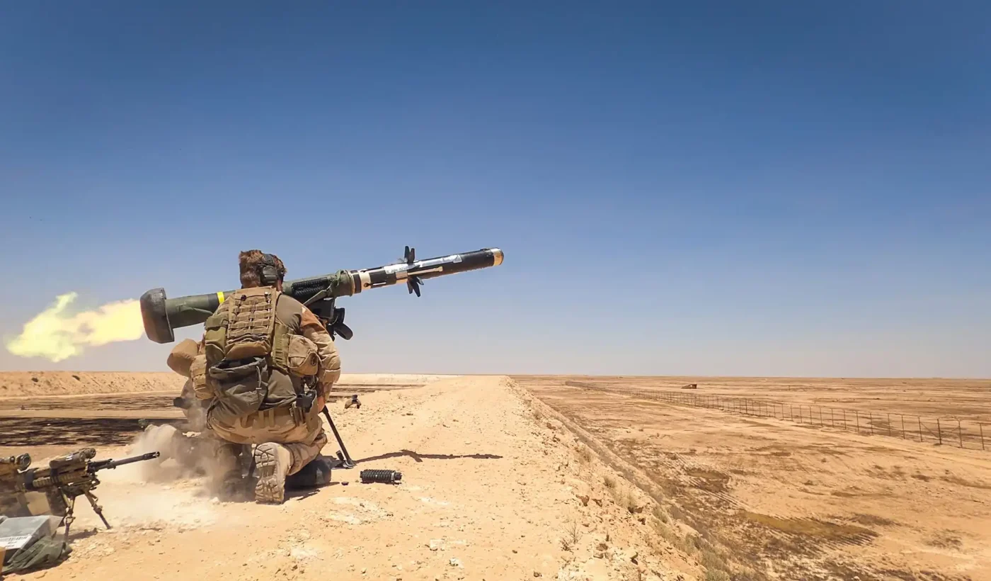 A soldier kneeling in the desert fires an FGM-148 Javelin anti-tank missile, with the back-blast visible