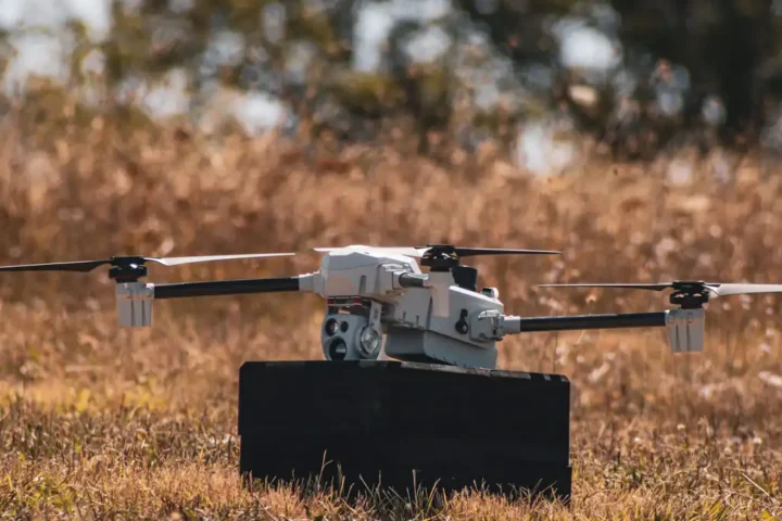A small white quadcopter-type loitering munition with a camera sensor, standing on a dark block in dry grass