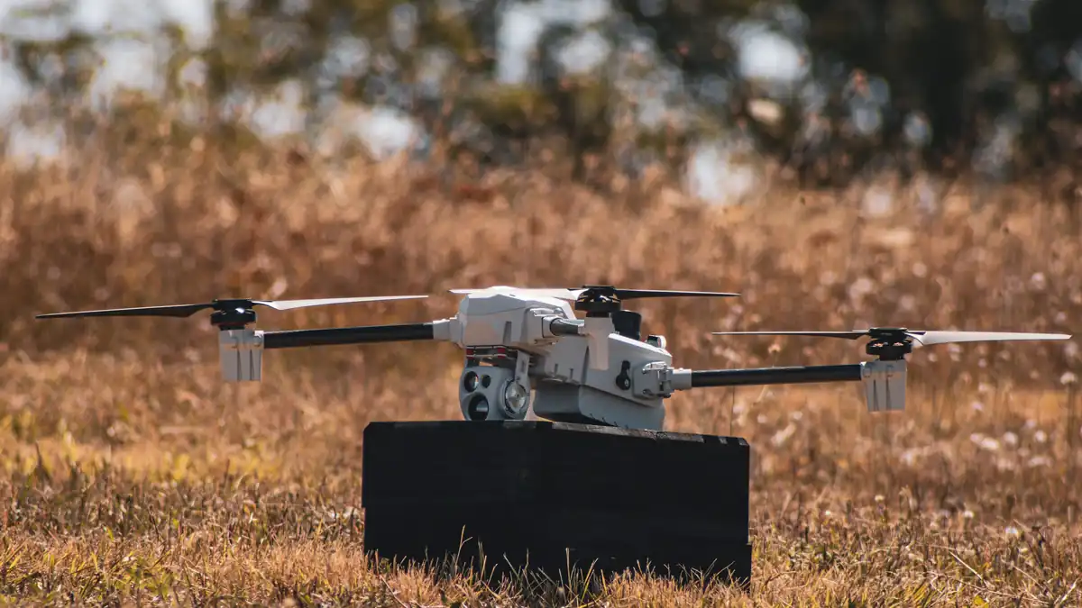 A small white quadcopter-type loitering munition with a camera sensor, standing on a dark block in dry grass