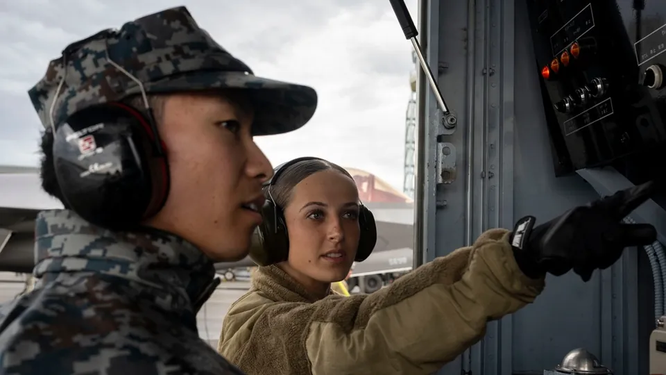 A U.S. Air Force airman and a Japan Air Self-Defense Force airman wearing ear protection at a refuelling station with fighter aircraft behind them