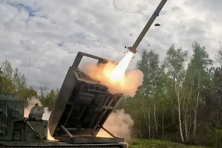 A rocket leaves the launcher of a tracked rocket artillery vehicle in a wooded training area under a cloudy sky