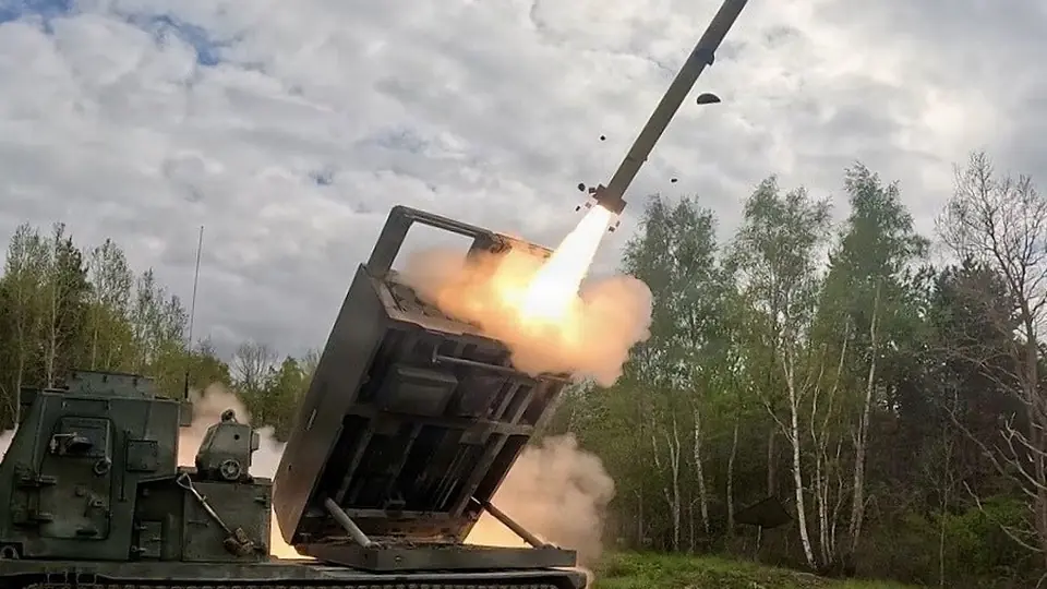 A rocket leaves the launcher of a tracked rocket artillery vehicle in a wooded training area under a cloudy sky