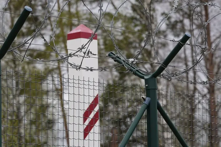 Green-posted wire-mesh fence topped with razor wire on the Latvian border, with a red-and-white border marker behind it