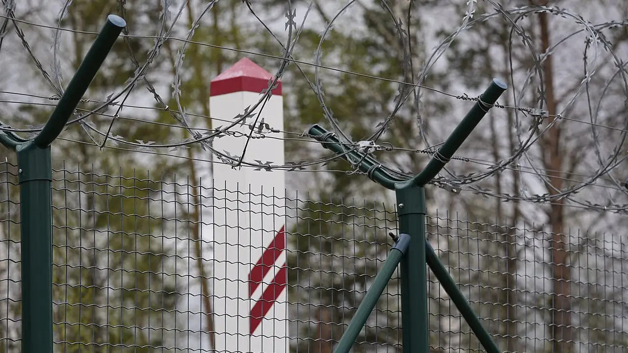 Green-posted wire-mesh fence topped with razor wire on the Latvian border, with a red-and-white border marker behind it