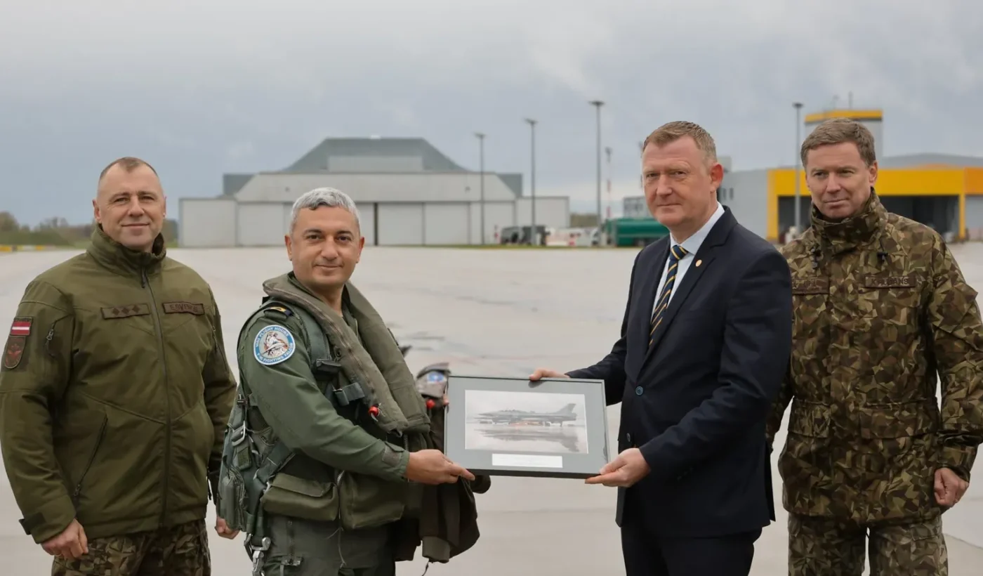 Four men on the apron at Lielvārde air base: a Turkish Air Force pilot in flight gear and Latvian Defence Minister Raivis Melnis in a dark suit hold a framed picture of a jet between two officers in uniform
