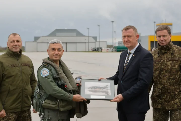 Four men on the apron at Lielvārde air base: a Turkish Air Force pilot in flight gear and Latvian Defence Minister Raivis Melnis in a dark suit hold a framed picture of a jet between two officers in uniform