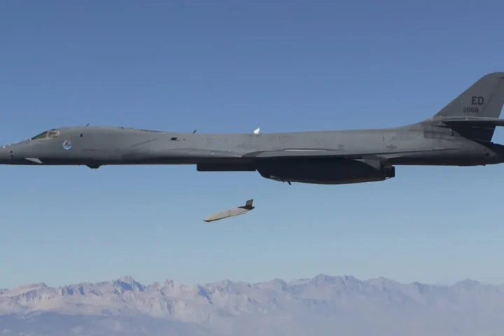 A B-1B Lancer bomber in flight over mountains with an LRASM anti-ship missile dropping away beneath its fuselage