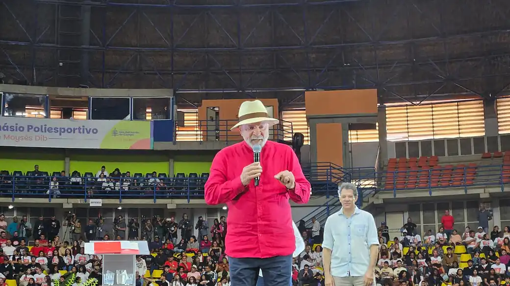 Lula da Silva speaking into a microphone at a political event in a sports hall, in a red shirt and straw hat