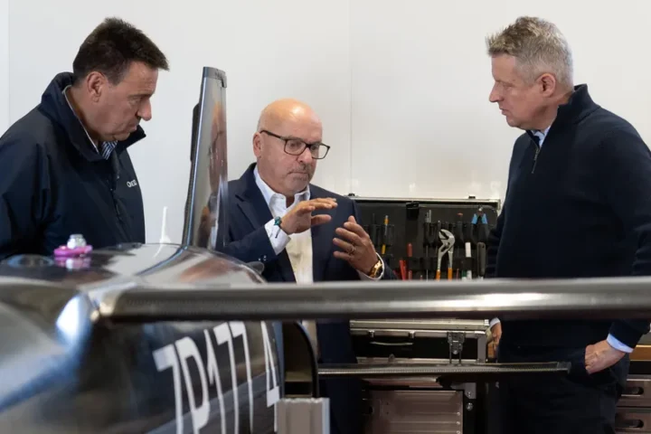 Three men in conversation beside a dark uncrewed aircraft in a workshop with tools on the wall