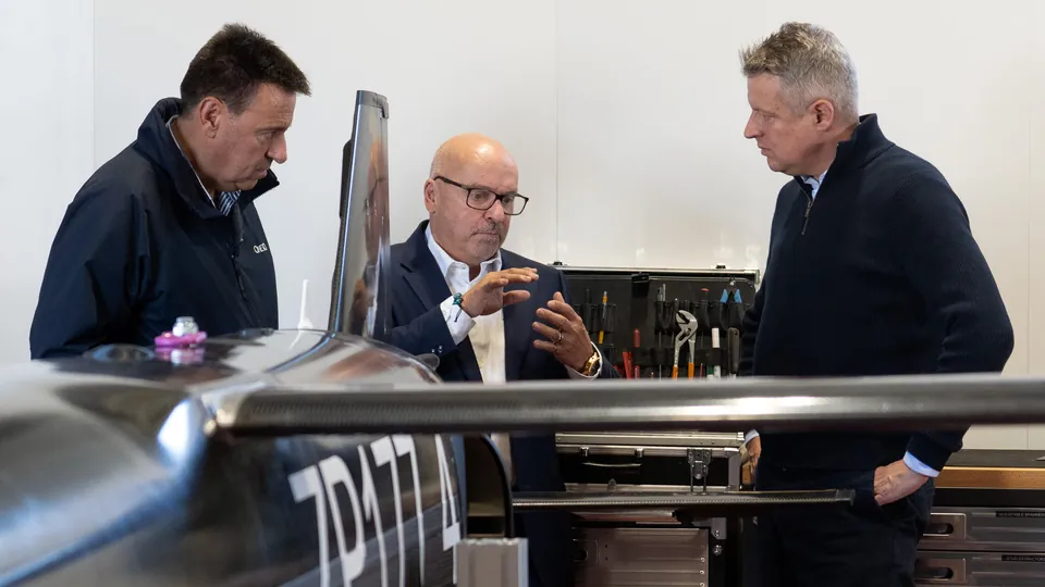 Three men in conversation beside a dark uncrewed aircraft in a workshop with tools on the wall