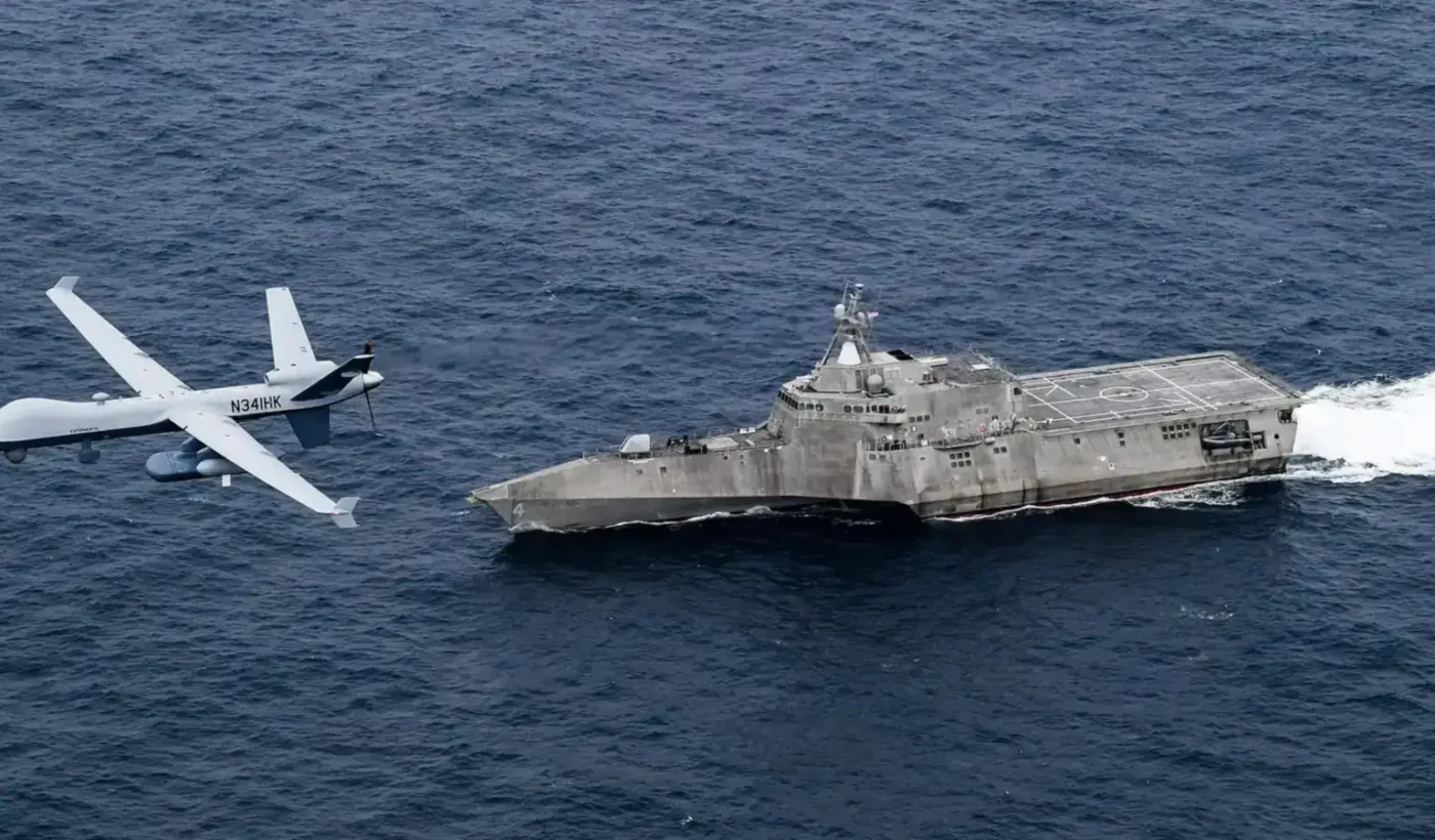 An MQ-9B SeaGuardian unmanned aircraft flies low over the sea next to the littoral combat ship USS Coronado