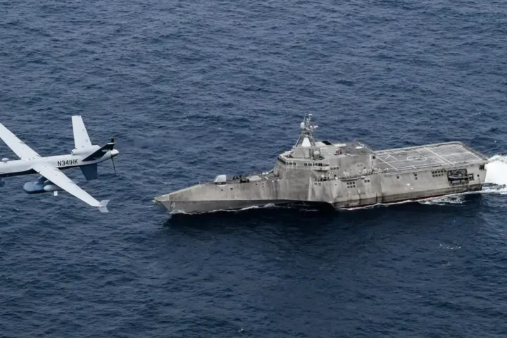 An MQ-9B SeaGuardian unmanned aircraft flies low over the sea next to the littoral combat ship USS Coronado