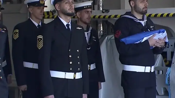 Hellenic Navy personnel in uniform line up on the frigate Nearchos while a sailor holds the folded Greek flag before it is raised