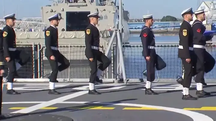 Hellenic Navy personnel march in formation on the flight deck of the frigate Nearchos, with the port of Lorient in the background