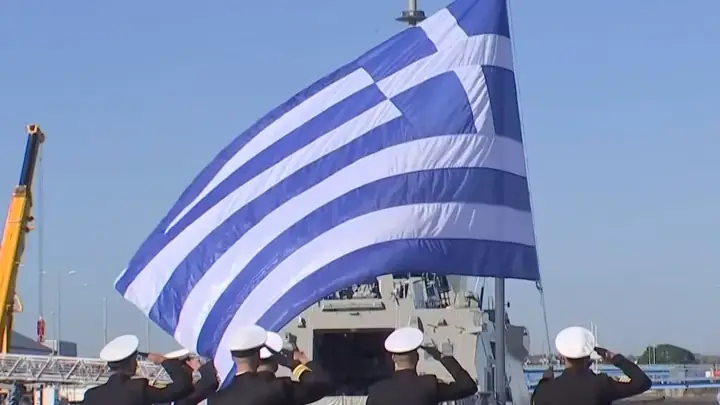 A large Greek flag flies from the mast of the frigate Nearchos as Hellenic Navy personnel salute in the port of Lorient