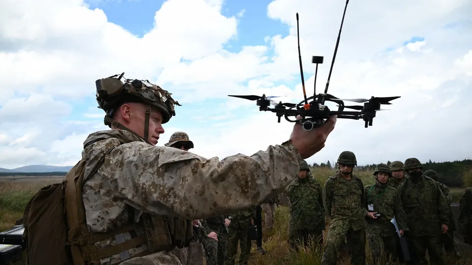 A U.S. Marine holds up a small quadcopter drone during a briefing at Camp Fuji, Japan, with soldiers in camouflage gathered behind him