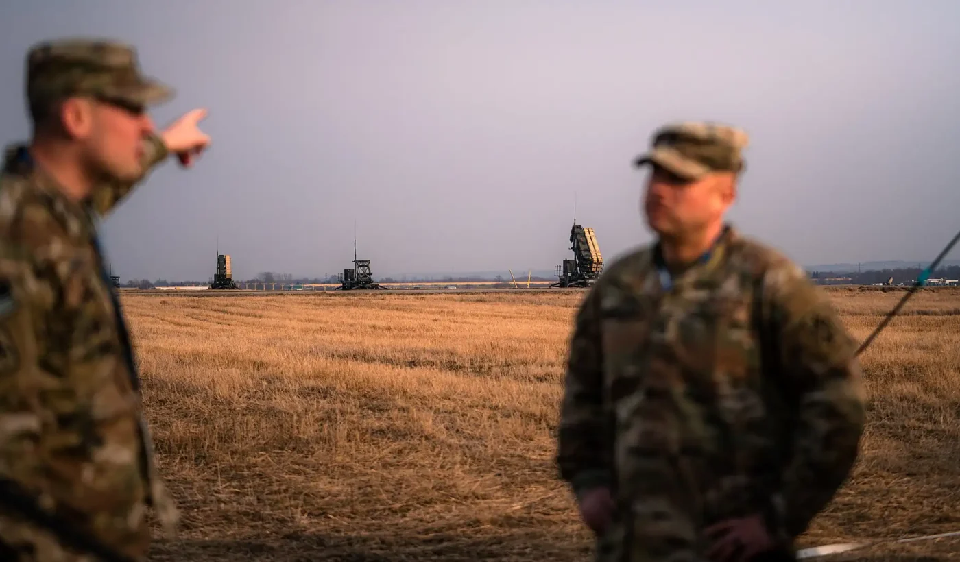 Patriot missile launchers and radar on an open field in Poland, with two blurred US Army soldiers in the foreground