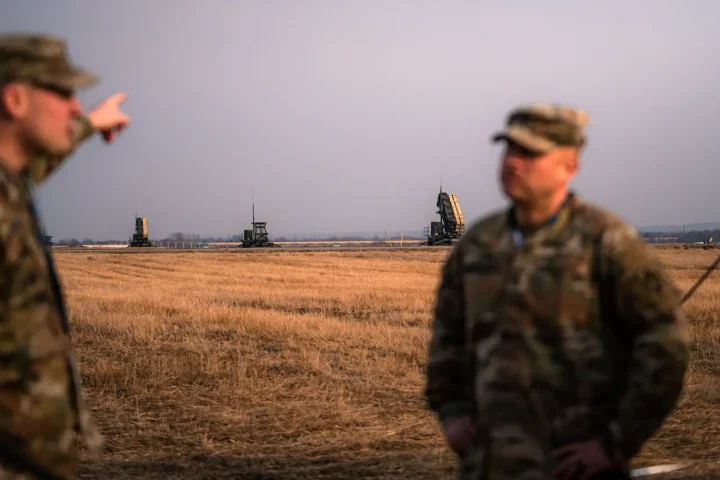 Patriot missile launchers and radar on an open field in Poland, with two blurred US Army soldiers in the foreground