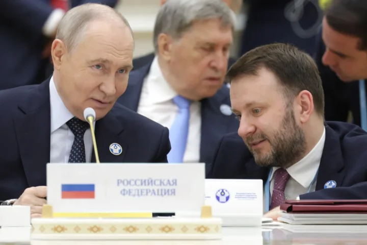 Vladimir Putin seated at the table at the CIS summit in Ashgabat, behind the nameplate of the Russian Federation, with an aide beside him