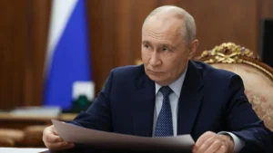 Vladimir Putin seated at a desk in the Kremlin with a Russian flag behind him, holding papers