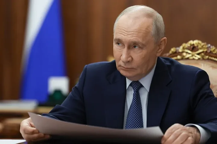 Vladimir Putin seated at a desk in the Kremlin with a Russian flag behind him, holding papers