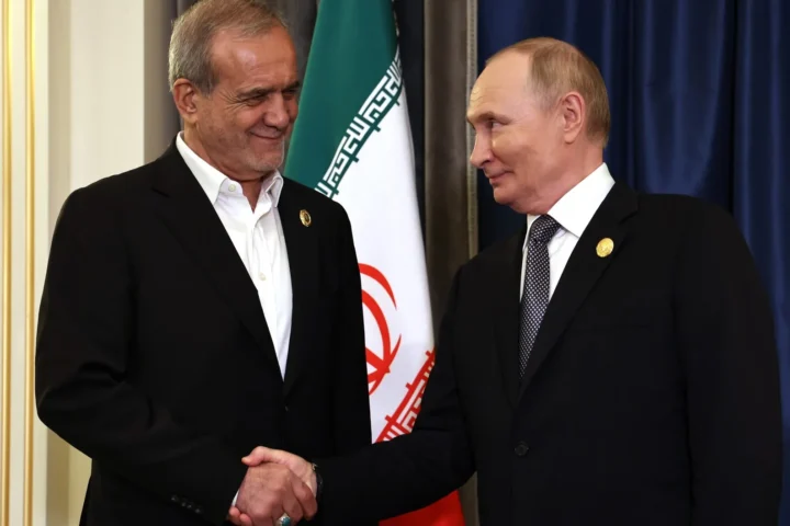 Vladimir Putin and Masoud Pezeshkian shaking hands in front of the flag of Iran